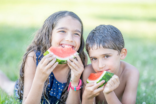 Two adorable siblings, a brother and sister are enjoying fresh watermelon outdoors on a summer day. They are at a water park and dressed in their bathing suit.