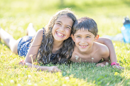 A happy brother and sister duo are lying down on green grass outdoors on a summer day. They are embracing each other. They are dressed in their bathing suits. They are smiling at the camera.