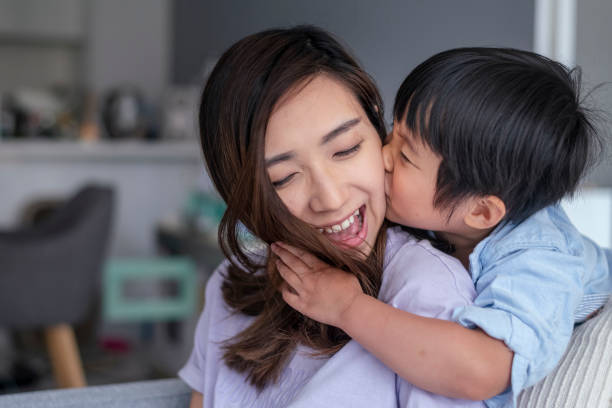 I love you Mommy! Close up shot of an adorable preschool age boy of Asian descent embracing his mom and giving her a kiss on the cheek. The loving mom is smiling with appreciation and affection. The family is spending quality time together at home. KISSING stock pictures, royalty-free photos & images