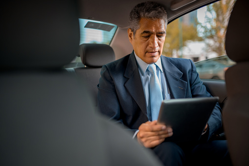 Senior businessman sitting on the back seat of the car and using digital tablet
