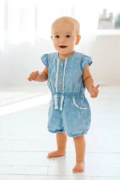 Photo of Very cute and funny baby in denim sarafan make first steps. Smiling little girl learning to walk. Concept of kids growth and happy childhood