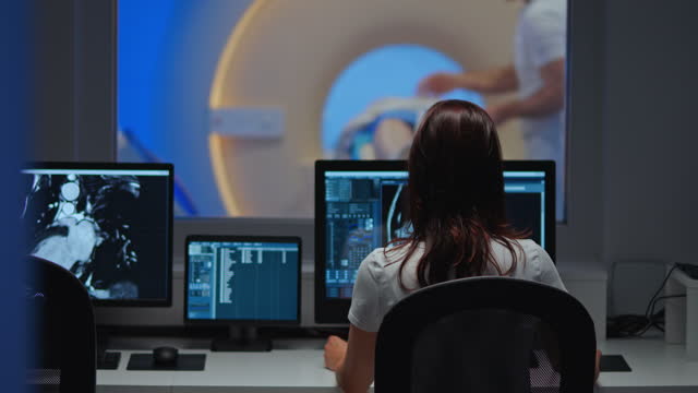 Wide dolly shot of a female radiology technologist sitting in the control room as the MRI technologist is preparing the patient in the imaging room. Shot in Slovenia.