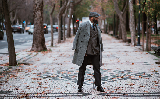 A dapper mature African senior with a black well-groomed beard is standing in the middle of an autumn boulevard and looking aside, with an elegant coat, hat, and a hand-made plaid suit on him
