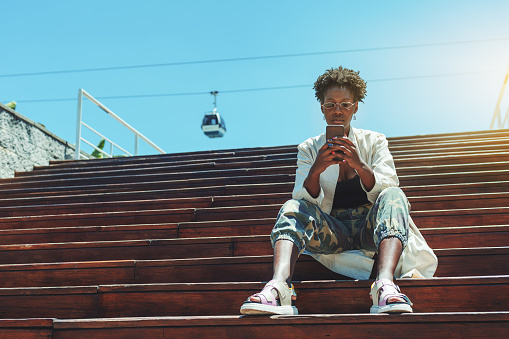 A fancy young African female tourist is phoning to see a list of attractions, while sitting on the wooden steps of outdoor stairs, with a ropeway cabin in front of the sky in a defocused background