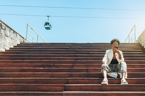 A fancy young African female is sitting on the wooden steps of the stair and using her smartphone to choose the best tourist route, a ropeway car in the background, a copy space place on the left