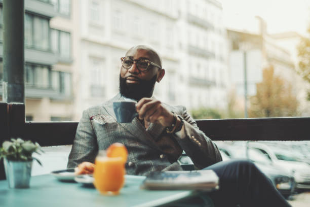 Black senior in a street restaurant The portrait of a handsome stylish wealthy African guy with a beautiful black beard, in glasses, bald, in an elegant suit, sitting on a rainy morning in a street cafe and drinking delicious coffee capital stock pictures, royalty-free photos & images
