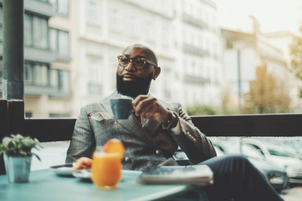 Black senior in a street restaurant The portrait of a handsome stylish wealthy African guy with a beautiful black beard, in glasses, bald, in an elegant suit, sitting on a rainy morning in a street cafe and drinking delicious coffee well dressed stock pictures, royalty-free photos & images