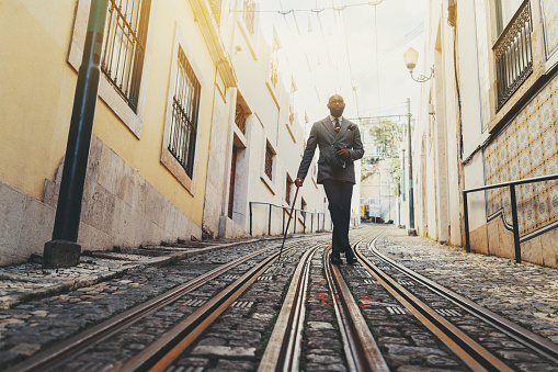 A dapper black senior man with a well-groomed beard and totally hairless is leaning against his fancy walking stick while standing on the tramway track of a narrow street of a European city