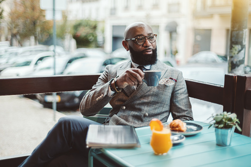 A fancy bearded hairless senior entrepreneur in glasses and a stylish custom-made costume, is sitting in an outdoor cafe in the morning and drinking a delicious coffee and pensively looking aside