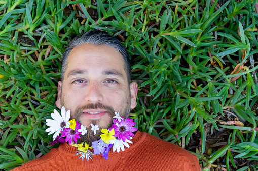 Handsome good-looking happy relaxed latin male colorful portrait, looking at camera with a beautiful smile and with multicolor spring flowers in his beard relaxing laying down at a green grass, wearing an orange sweater.

Spring Concept. Springtime daisies Concept. Space for copy text.