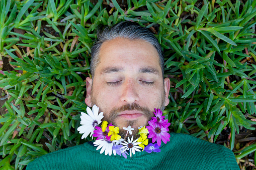 Handsome good-looking happy relaxed latin male colorful portrait, in front of the camera with his eyes closed and a multicolor spring flowers in his beard relaxing laying down at a green grass, wearing a green sweater.

Spring Concept. Springtime daisies Concept. Space for copy text.