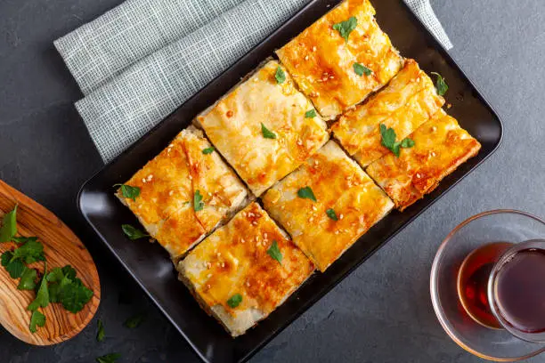 Turkish kiymali borek served on a tray after being sliced into square shapes. Fresh parsley leaves are sprinkled on top for added flavor. A glass of Turkish tea in special tea glass nearby.