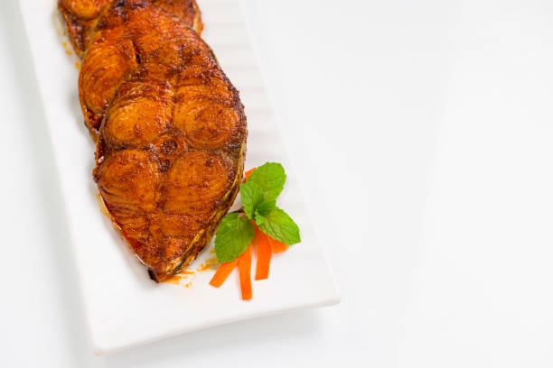 Seer fish fry arranged beautifully and garnished with onion, lemon and tomato slices on white rectangle shape ceramic plate placed on white textured background.