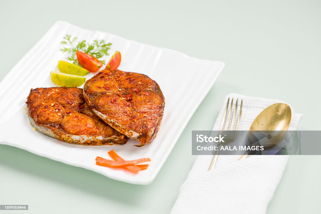 Seer fish fry Seer fish fry arranged beautifully and garnished with onion, lemon and tomato slices on white square shape ceramic plate placed on ice  blue textured background with serving spoon placed on a white napkin. Opah Stock Photo