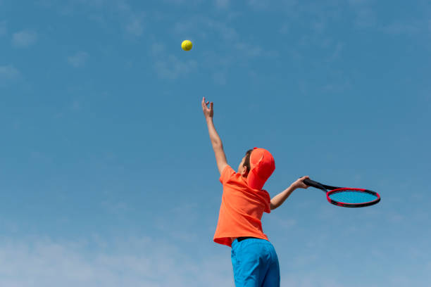 오렌지 스포츠웨어를 입은 어린이 소년 테니스 선수, 라켓을 입고 서브 볼을 연주하는 법을 배움. 키즈 스포츠 테니스 게임, 학교 또는 클럽에서 훈련. 행동에 아이 운동 선수. 배경 복사 공간 - 주니어 레벨 뉴스 사진 이미지