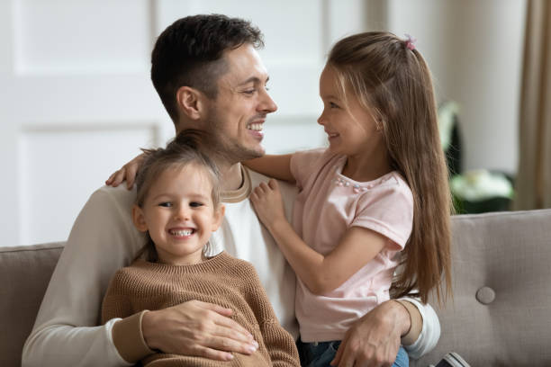 istock Father sitting on couch hugging two little daughters 1309383524