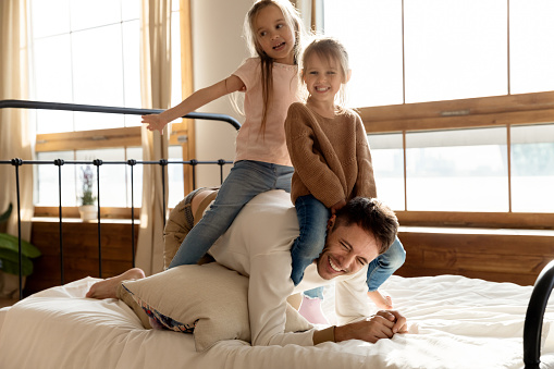 In bedroom two little cheery daughters play with dad, saddled him seated on his back. Laughing father giving piggy back ride to children enjoy funny activity in morning at home. Family games concept