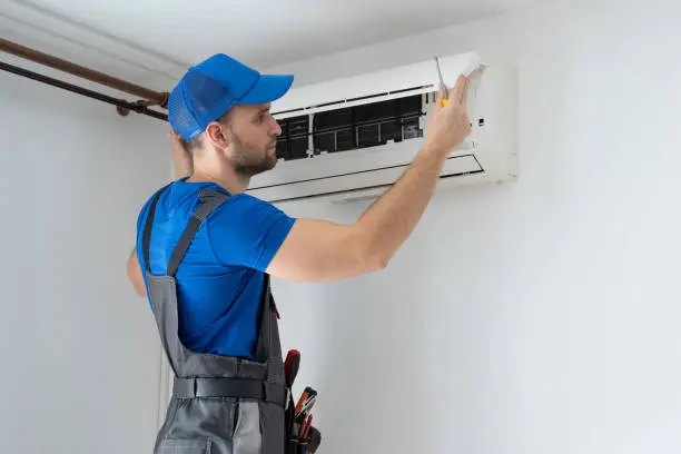 Male technician in overalls and a blue cap repairs an air conditioner on the wall.