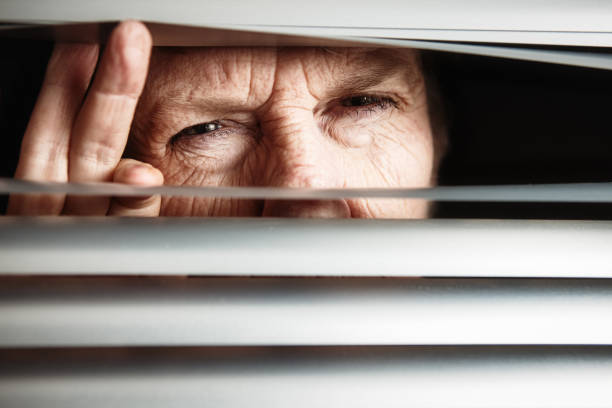 Elderly woman peering anxiously through window blinds Senior woman stares out suspiciously through her blinds, her fingers separating them. squint eyes stock pictures, royalty-free photos & images