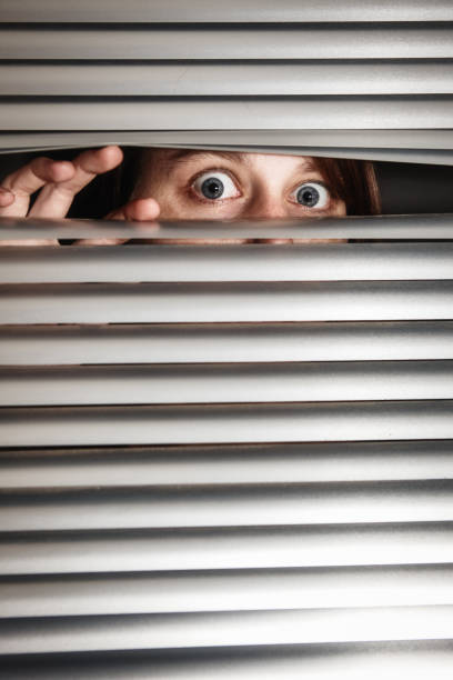 istock Young woman's eyes peering anxiously through window blinds 1309388217