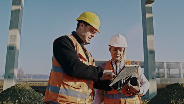 建設現場での建物の建設進捗状況を評価するためのデジタルタブレットを使用した建築請負業者とエンジニアのスローモーションショット。日の出に撮影されたショット。8K解像度で撮影。