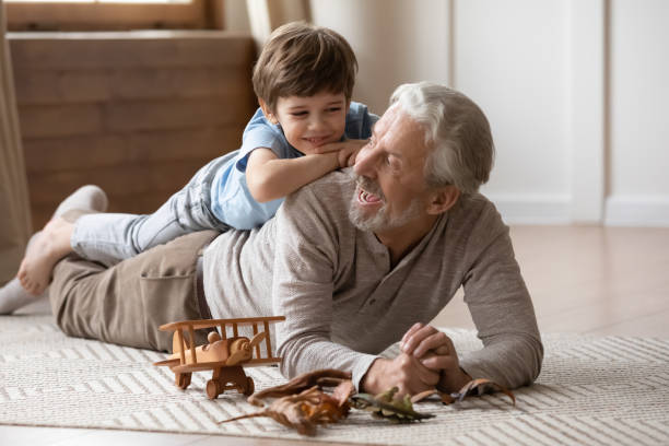 Overjoyed mature grandfather playing with adorable grandson at home Overjoyed mature grandfather playing with adorable grandson at home, older man lying on warm floor with cute little boy grandchild on back, family spending leisure time together, having fun young hot stock pictures, royalty-free photos & images