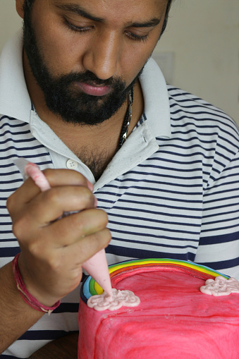 Stock photo showing an Indian man decorating a cake with a icing /piping bag. Circular homemade chocolate cake, with butter icing cream between layers of sponge. The top of the cake has been covered with red fondant icing and decorated with icing rainbow and clouds.