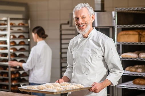 Working mood. Adult gray-haired happy man in uniform with tray of bagels and woman behind near rack of bread working in bakery