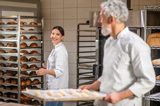 Bakery, colleagues. Joyful woman in white uniform near rack of buns and turned gray-haired employee with tray of croissants at bakery