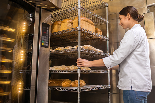 Baking bread. Satisfied woman in white jacket standing near oven in bakery watching baking in good mood