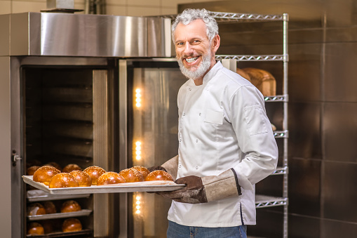 Hot buns. Joyful gray-haired bearded man in gloves with baking sheet of freshly baked buns near oven in bakery