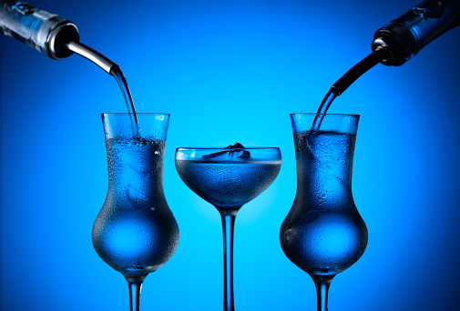 Vodka is poured from bottles into glasses with ice. Blue background.