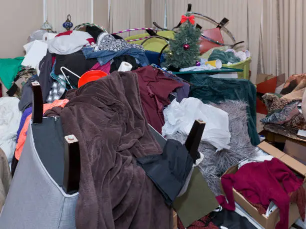 Side view of a large pile of household objects stacked in the middle of a room including a chair, clothes, papers, blankets, bowls, cups, kitchen items, Christmas tree and decorations.