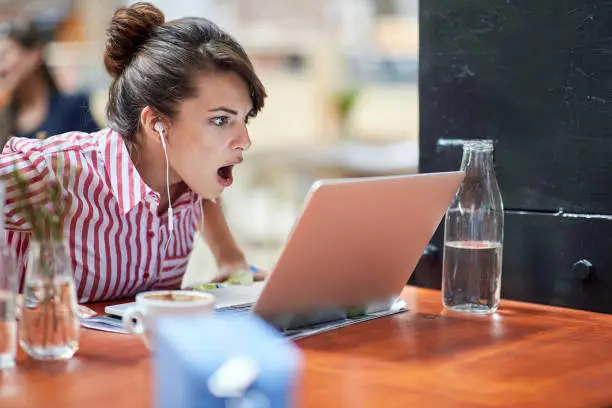 Photo of amazed young female watching content on her laptop with headphones in ears, can't believe what she saw, with her mouth open. amazed, astonished reaction concept