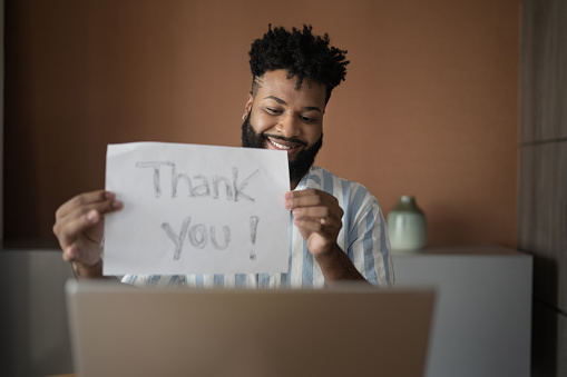 Young man holding paper with 'thank you' text on a video call at laptop at home
