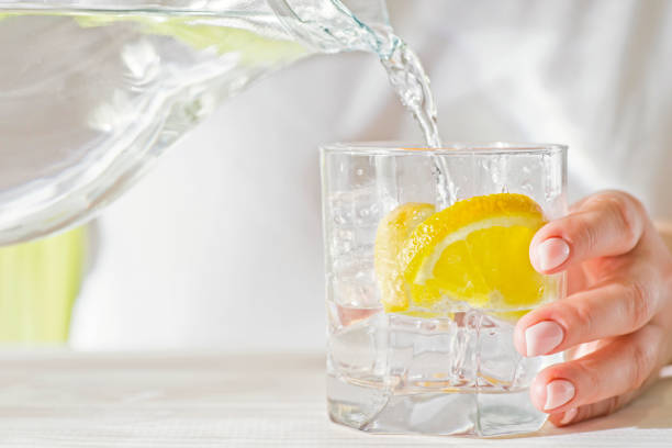 mani femminili che versano acqua dal decanter in un bicchiere con limone e ghiaccio. concetto di salute e dieta. dissetarsi in una giornata calda. - juice glass healthy eating healthy lifestyle foto e immagini stock