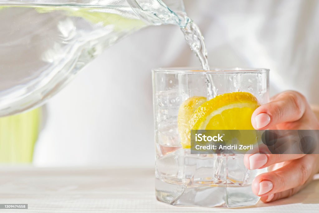 Female hands pouring water from the decanter into a glass beaker with lemon and ice. Health and diet concept. Quenching thirst on a hot day. Water Stock Photo