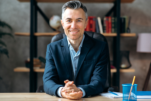 Portrait of an attractive, successful, influential, adult caucasian manager, businessman or lawyer in stylish formal suit, sitting at work desk in the office, looks at the camera friendly smiling