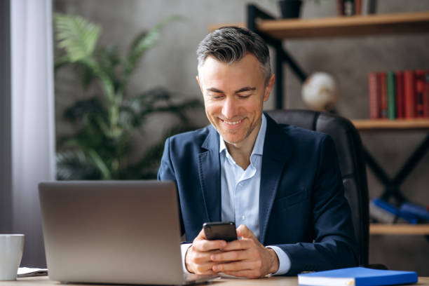 istock Online chat. A cheerful, handsome caucasian businessman is sitting at his desk, using a mobile phone while working, browsing the Internet, answering email, and texting with a friend or family, smiling 1309478246