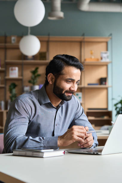 homme d’affaires indien barbu de sourire travaillant sur l’ordinateur portatif au bureau à la maison. jeune étudiant indien utilisant l’étude à distance d’ordinateur, formation virtuelle sur la réunion d’appel vidéo, regardant le webinaire  - learning male studying smiling photos et images de collection