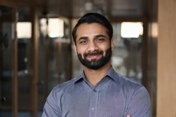 Photo of Portrait of young happy indian business man executive looking at camera. Eastern male professional teacher, smiling ethnic bearded entrepreneur or manager posing in office, close up face headshot.