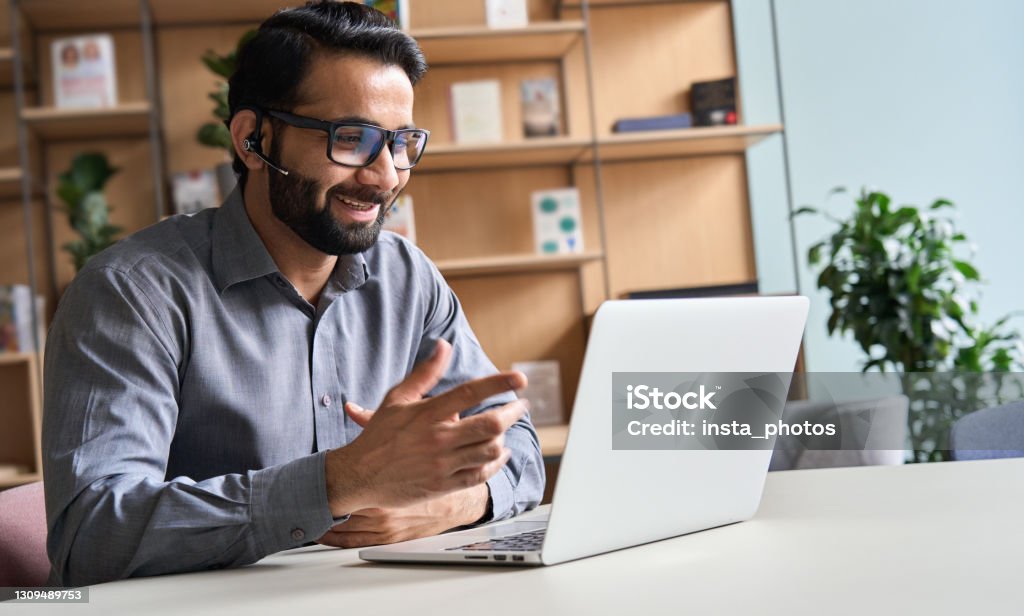 Happy indian business man remote teacher, customer support manager wearing headset talking at virtual meeting consulting client on video call giving distance learning class at home office call center. Using Phone Stock Photo