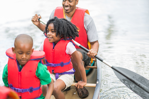 A beautiful young family with two children are enjoying a canoe trip together. The children are seated in the middle of the canoe. The focus of the image is on the young girl who is seated in the middle of the canoe with his brother.