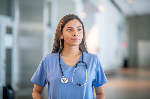 A young female medical student is standing inside a medical building. She is wearing blue medical scrubs and is looking away from the camera.