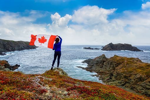 Adventurous woman holding a Canadian Flag on a Rocky Atlantic Ocean Coast during a cloudy and blue sky day. Taken in Sleepy Cove, Crow Head, Twillingate, Newfoundland, Canada.