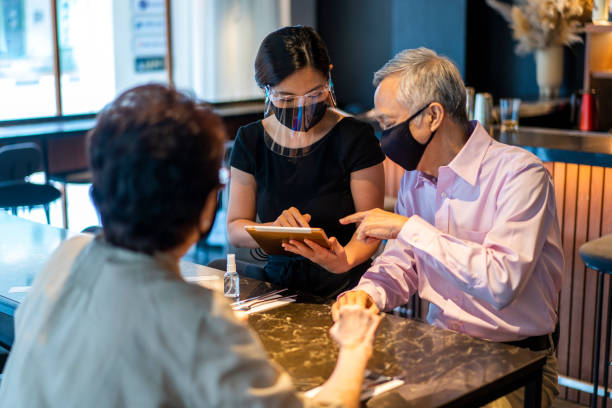 istock Asian waitress with face mask and face shield taking food orders on her digital tablet, from senior couple customers. 1309505559