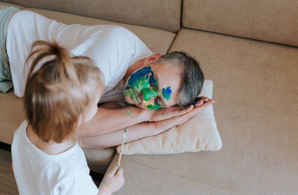uma garotinha pinta o rosto de seu pai com um pincel e aquarelas enquanto ele dorme no sofá - child art paint humor - fotografias e filmes do acervo
