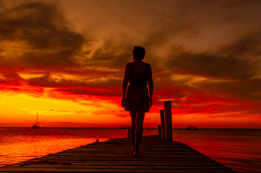 A young woman walking towards the Caribbean Sea in the orange sunset at the West End beach of Roatan. Honduras