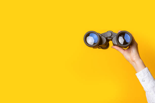 mano femenina sostiene binoculares negros sobre un fondo amarillo brillante - binaculars fotografías e imágenes de stock