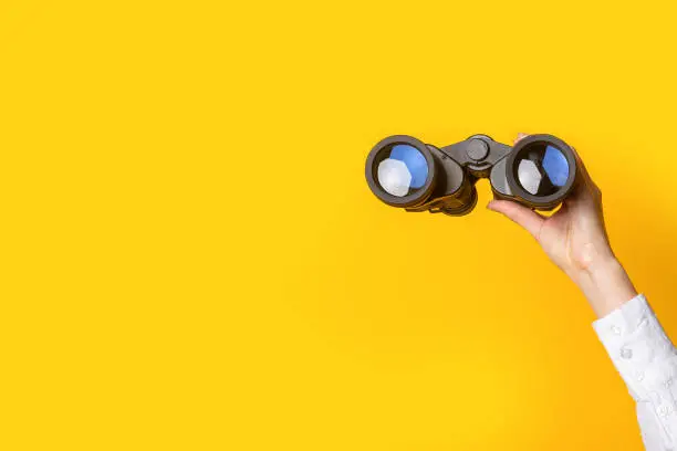 Photo of female hand holds black binoculars on a bright yellow background